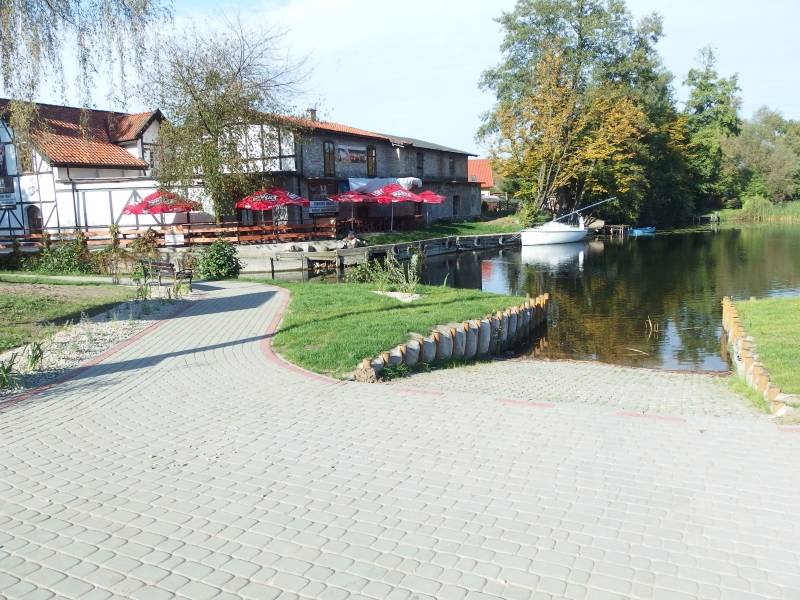 Przystań wodna w Miłomłynie. Na pierwszym planie slip. W głębi za kanałem restauracja i miejcsa pod parasolami.