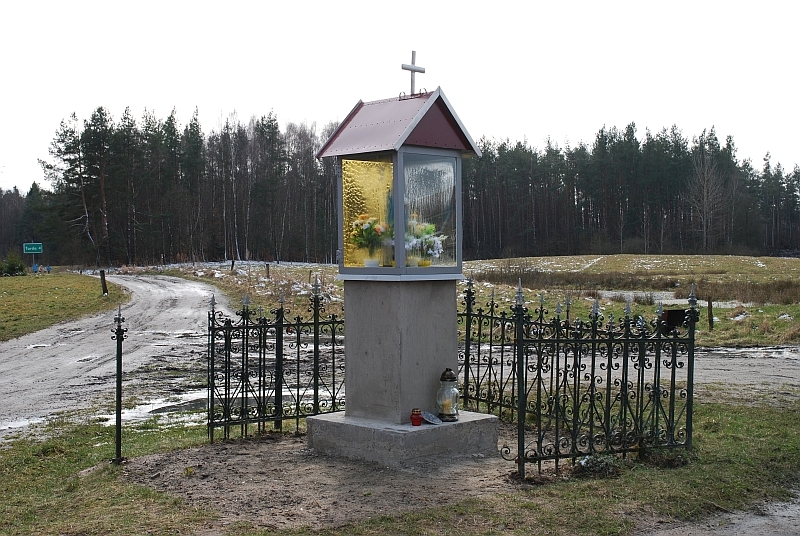 Kapliczka ze szklaną witryną w ogrodzeniu z metaloplastyki. Krajobraz wczesnej wiosny